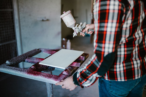 Young man painting a wood board in his workshop using a spray gun.