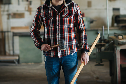 Carpenter wearing a plaid shirt standing in his workshop and holding a hammer and a piece of wood.